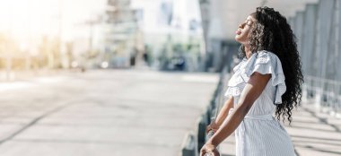 Uzun, kıvırcık siyah saçlı Afro-Amerikalı genç kadın şehir kaldırımında duruyor, güneşin ve temiz havanın tadını çıkarıyor. Beyaz, çizgili kollu bir elbise giyiyor, fotokopi odası.