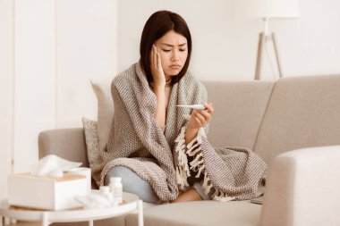 Battaniyeye sarılı bir kadın kanepede oturuyor, termometreyle ateşini ölçerken endişeli görünüyor..