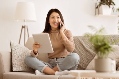 Bir kadın kanepede oturuyor, tablet kullanırken gülümsüyor ve telefonla konuşuyor..