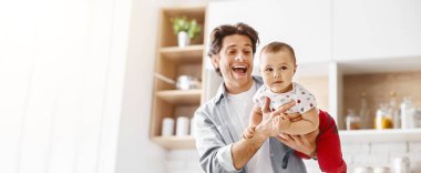 Görüntü, bebeğini mutfakta tutan mutlu bir babayı gösteriyor. Baba gülümsüyor ve kameraya bakıyor. Bebek de kamera dışı bir şeye bakıyor.