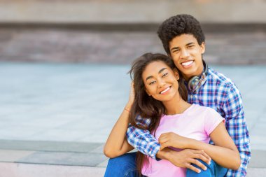 Dışarıdaki merdivenlerde oturan siyah bir çift kameraya gülümsüyor. Adam ekose gömlek ve kot pantolon giyerken kız pembe gömlek ve kot pantolon giyiyor..