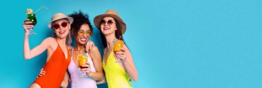 Mayo, şapka ve güneş gözlüğü takan üç arkadaş mavi bir arkaplan önünde kokteyl içerken gülümsüyor. Soldaki kadın elinde pipetle yeşil bir kokteyl tutuyor.