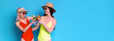 İki mayo giymiş kadın gülümsüyor ve parlak mavi bir arka planda kokteyllerle kadeh kaldırıyorlar. Bir kadın kırmızı mayo ve güneşlik giyerken, diğeri sarı mayo giyiyor.