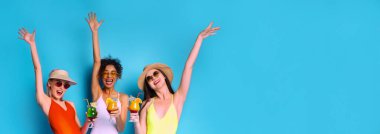 Mayo ve güneş gözlüğü takan üç kadın mavi arka planda yan yana duruyorlar. Kollarını havaya kaldırdılar ve her kadın bir kokteyl içeceği tutuyor.