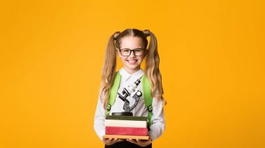 Gülümseyen Schoolgirl Holding Kitap Yığını Ve Mikroskop Sarı Stüdyo Arka Plan. Okul Konseptine geri dönelim. Boş Alan