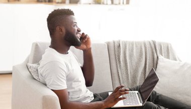 Evinden çalışan siyahi bir girişimci, kucağında bilgisayarla koltukta oturuyor, telefonla konuşuyor, yan gözle bakıyor.