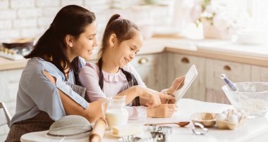 Bir anne ve küçük kızı modern mutfaklarında tablete bakıyorlar. İkisi de gülümsüyor ve birlikte bir pişirme projesinde çalışıyor gibi görünüyorlar.