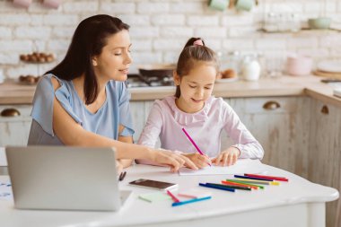 Bir anne, genç kızıyla mutfak masasında oturur. Kız, pembe pastel boyayla bir kağıt parçasına boya sürerken anne de kağıdı izleyip işaret ediyor..