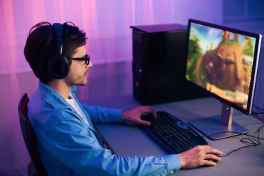Kulaklık ve gözlük takan genç bir adam bilgisayar monitörünün önünde oturmuş video oyunu oynuyor. Oda mor ışıklarla aydınlanıyor, canlı ve enerjik bir atmosfer yaratıyor..
