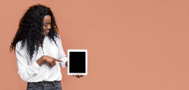 Kıvırcık siyah saçlı, gülümseyen siyah bir kadın beyaz düğmeli gömlek ve koyu mavi kot pantolon giyiyor. Sol elinde bir tablet tutuyor ve sağ işaret parmağıyla ekranı işaret ediyor.