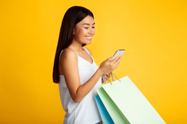Beyaz atletli gülümseyen Asyalı genç bir kadın sarı arka planda duruyor. Sağ elinde akıllı bir telefon, sol elinde de alışveriş torbaları var.
