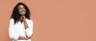 Siyah kıvırcık saçlı, gülümseyen Afro-Amerikalı genç bir kadın. O, beyaz bir düğmeli gömlek giyiyor. Kadın yukarı bakıyor, çenesini elinin üzerinde dinlendiriyor ve gülümsüyor. Arka plan açık turuncu.