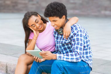 Genç siyah bir çift dışarıda basamaklarda oturuyor. Kız pembe bir gömlek giyiyor ve kolunu erkeklerin omzuna dolamış. Mavi bir ekose gömlek giyiyor ve bir tablete bakıyor..