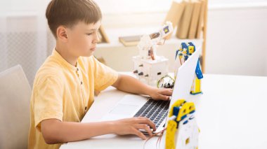 Genç bir çocuk dizüstü bilgisayar kullanarak robot bilimi öğreniyor. Çocuk bir masada oturuyor ve dizüstü bilgisayarın ekranına bakıyor. Bilgisayarın arkasında bir robot kol var..