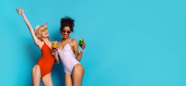 İki kadın parlak mavi bir arka planın önünde duruyorlar, gülümsüyorlar ve ellerinde kokteyller var. Kadınlar mutlu ve kaygısızdır. Harika vakit geçiriyor gibi görünüyorlar, kopyalama uzayı.