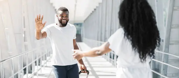 Stock image A smiling black man with a beard is standing in an airport terminal, waving hello to a woman walking towards him. He is holding a suitcase and looks happy to see her.