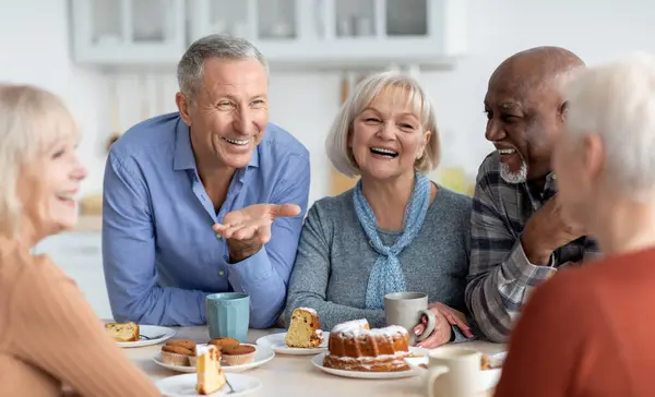 stock image Multiracial group of happy senior people sitting around table drinking tea with cake and having conversation, smiling and laughing, having home party or enjoying time at nursing home