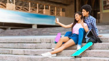 Genç siyah bir çift önlerinde kaykaylarla merdivenlerde oturuyor. Kız selfie çekerken oğlan barış işareti yapıyor. İkisi de gülümsüyor ve birbirlerine eşlik etmekten zevk alıyorlar..