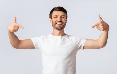 Beyaz tişörtlü bir adam kolları açık beyaz bir arka plana karşı duruyor, yüzünde kocaman bir gülümsemeyle kendini işaret ediyor..