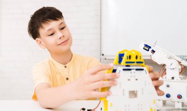 Kısa kahverengi saçlı genç bir çocuk bir masada oturuyor ve bir robot yapıyor. Sarı bir gömlek giyiyor ve robot üzerinde çalışırken yüzünde konsantre bir ifade var.