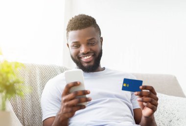 Afro-Amerikalı bir adam parlak beyaz bir geçmişi olan bir koltukta oturuyor, bir elinde akıllı telefonu, diğer elinde bir kredi kartı ile online alışveriş yaparken gülümsüyor..