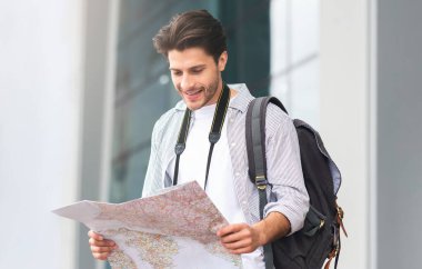 Mutlu Turistler. Ellerinde şehir haritasıyla havaalanının yanında duran gülümseyen genç adam, rotasını kontrol ediyor, boş uzay.