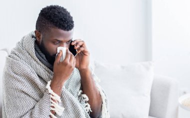 Battaniyeye sarılı siyah bir adam kanepede oturur ve burnunu silerken telefonla konuşur. O açıkça hasta. Arka planda beyaz bir duvar ve beyaz bir kanepe olduğu için evinde gibi görünüyor..