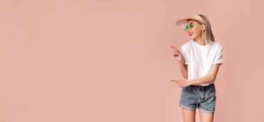 Güneş gözlüklü ve şapkalı bir kadın pembe bir arka planın önünde duruyor. Gülümsüyor ve sağ eliyle solu işaret ediyor, kopyalama alanı.