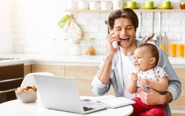 Bir baba önünde dizüstü bilgisayarıyla mutfak masasında oturuyor. O, telefonda konuşuyor ve bebeğini tutarken gülümsüyor. Bebek de gülümsüyor ve fotokopi odasına bakıyor.
