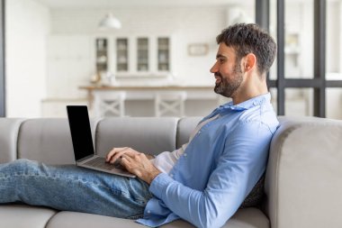 Bu resim gri bir kanepede dizüstü bilgisayar kullanan bir adamı gösteriyor. Kot pantolon ve açık mavi düğmeli bir gömlek giyiyor ve yazarken laptop ekranına bakıyor..