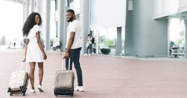 Havaalanında bavullarıyla yürüyen Afro-Amerikalı bir çift. Onların her ikisi de bagajlarını arkalarına çekiyor. Havaalanı geniş pencereli, kopyalama alanı olan modern bir bina.