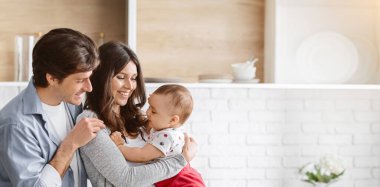 Bu resim modern bir mutfakta üç kişilik mutlu bir aileyi gösteriyor. Aileler bebeklerini kucağında tutarken gülümsüyor. O da yan tarafa bakıyor, boşluğu kopyalıyor.