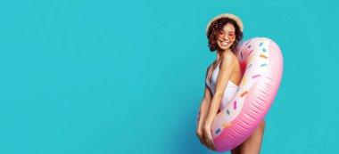 Beyaz mayo ve hasır şapkalı genç siyah bir kadın elinde pembe donut şekilli şişme bir havuz tutarken gülümsüyor parlak mavi bir arkaplan önünde.