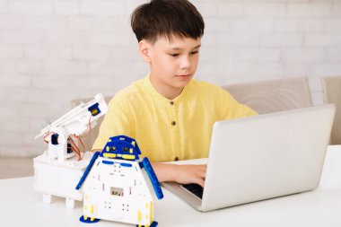 Genç bir çocuk masada oturuyor, küçük bir robotu programlamak için dizüstü bilgisayar kullanıyor. Robot mavi aksanlı beyaz plastikten yapılmış ve robotik kolu var. Çocuk ekrana odaklanmış.
