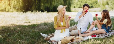 Kırsal kesimde piknik yapıp çimenlerde elma yiyen genç aile