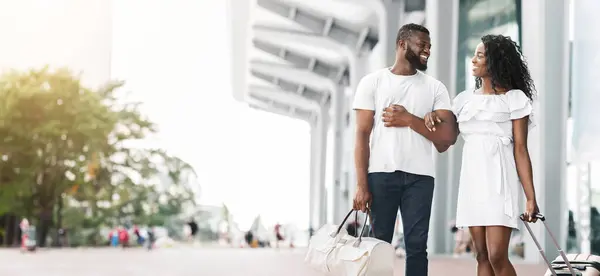 stock image A smiling African American couple walks together outside a modern building on a sunny day. The man has his arm around the womans waist and they are both carrying luggage, copy space