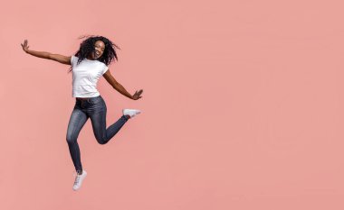 Bu resim siyah saçlı, beyaz tişörtlü ve kot pantolonlu, pembe arka planda zıplayan genç bir Afro-Amerikan kadını gösteriyor. Gülümsüyor ve eğleniyor gibi görünüyor..