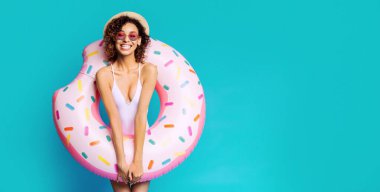 Beyaz mayo ve güneş gözlüğü takan gülümseyen siyah bir kadın büyük pembe renkli bir donut havuzu tutuyor. O, parlak mavi bir arka planın önünde duruyor..