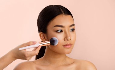 Görünmeyen bir el tarafından yüzüne makyaj yaptıran Asyalı bir kadın. Makyaj fırçası sağ yanağına sürerken kadın kameraya tarafsız bir ifadeyle bakıyor..