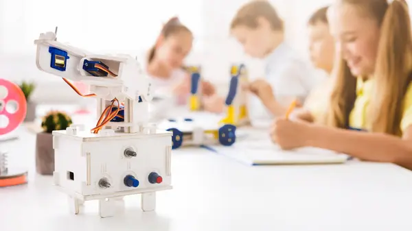 stock image A close-up shot of a white robotic arm on a table in a classroom setting. The robot is in focus and there are blurred children in the background who are building or working on projects.