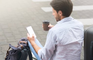 Mekik Otobüsü Bekliyor. Genç adam kahve içiyor ve akıllı telefon kullanıyor havaalanı binasının yanında otururken, Omuz manzaralı, bedava uzay manzaralı.