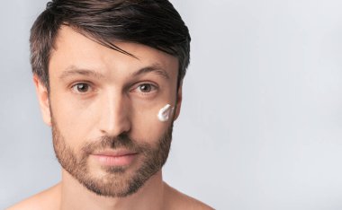 Sakallı bir adam kameraya bakarken yanağına krema sürer..