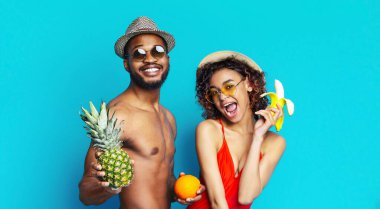 Siyah adam ve kadın gülümsüyor ve mavi arka planda kameraya poz veriyorlar. Adam güneş gözlüğü ve şapka takıyor ve ananas tutuyor. Kadın elinde muz ve portakal tutuyor..