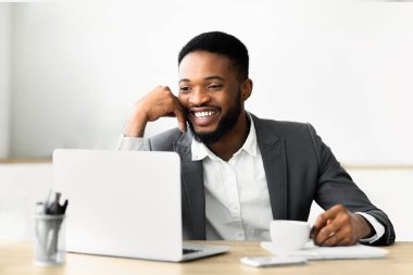 Takım elbiseli bir adam masasında oturmuş video görüşmesi sırasında dizüstü bilgisayara bakarken gülümsüyor. Önündeki masada bir fincan kahve ve bir telefon var..