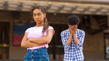 Genç siyah bir kız okul binasının önünde kollarını kavuşturmuş ve sert bir ifadeyle duruyor. Arka planda, yüzü ellerinde ağlayan genç bir çocuk vardı..