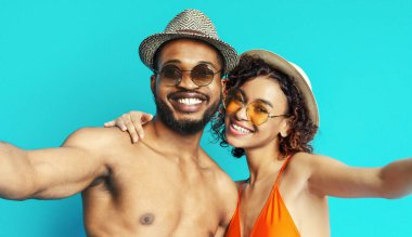 Her ikisi de güneş gözlüğü ve şapka takan gülümseyen Afro-Amerikan bir çift, parlak mavi bir arka planda selfie için birlikte poz veriyorlar. Belli ki yaz sıcağında birlikte vakit geçirmekten zevk alıyorlar..