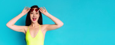 Sarı mayo giymiş genç bir kadın kalp şeklinde güneş gözlüğünü yüzüne tutarak geniş bir gülümseme sergiliyor. O, mavi bir arkaplanın önünde. Siyah saçları uzun ve düz, kopyalama alanı var.