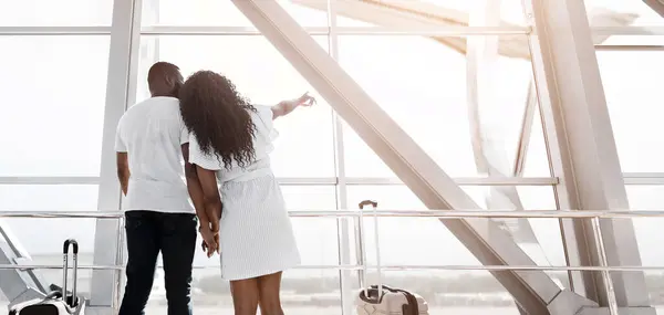 stock image African American couple stands near an airport window, gazing out at the view, while holding hands and waiting for their flight. The woman is pointing at something out of frame, copy space