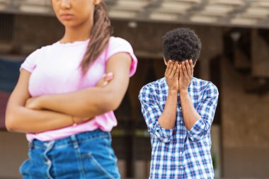 Saçları afro saçlı, genç bir siyah adam, yüzünü elleriyle kapatıyor, üzgün ya da utanmış görünüyor. Arkasında, genç bir kız kollarını kavuşturup başka tarafa bakıyordu.