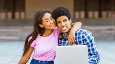 Genç siyah bir çift, pembe gömlek giyen uzun siyah saçlı bir kız ve ekose gömlek giyen kısa siyah saçlı bir adam kameraya gülümsüyorlar. Dışarıda dizüstü bilgisayarla oturuyorlar.